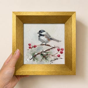 May include: An oil painting of a black-capped chickadee perched on a snowy branch with red berries, framed in a square gold-coloured frame. The bird has a black head and white cheeks, and the painting features visible brushstrokes and a signature.