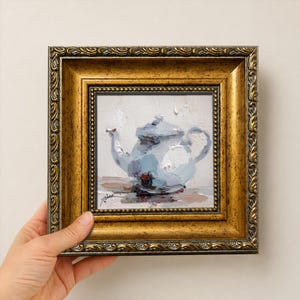 Puede incluir: Una pintura al óleo sobre lienzo que representa una tetera de color azul claro con pinceladas visibles, sobre un fondo neutro. La obra está enmarcada en un marco dorado metálico ornamentado con un borde interior de cuentas y motivos de hojas.
