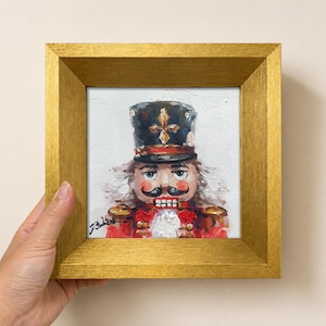 May include: A square oil painting of a nutcracker in a gold frame. The nutcracker wears a black hat with a gold emblem, a red uniform with gold details, and has a white beard. The painting features visible brushstrokes and a signature.