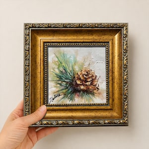 以下が含まれることがあります： 緑の松葉がついた茶色の松ぼっくりを描いた油絵で、ビーズ状の縁取りがある正方形の装飾的なアンティークゴールドの額縁に入っています。キャンバスの左下隅には筆致と署名がはっきりと見えます。