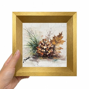 May include: A square oil painting in a gold frame featuring a textured depiction of a brown pinecone, a green pine sprig, and a dried oak leaf against a light cream background. The artist's signature is visible in black in the lower left corner.