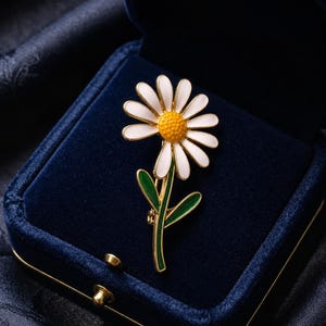 May include: A daisy flower brooch with white petals and a yellow center, set in a gold-tone metal frame. The stem and leaves are green. The brooch is displayed in a dark blue velvet jewelry box.
