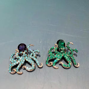 May include: Two octopus brooches. One is teal with a dark purple gem, the other is green with a green gem. Both have gold-coloured metal accents and are covered in small, sparkling crystals.
