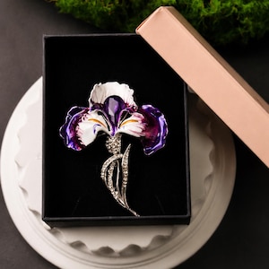Puede incluir: Un broche de flor de iris en tono plateado con pétalos de esmalte blanco y morado oscuro, con una piedra central morada facetada. El tallo y las hojas están adornados con cristales transparentes, presentado en una caja negra con tapa rosa claro.