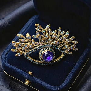 May include: A gold-toned eye-shaped brooch with a large purple center stone and blue and iridescent accents. The brooch is set in a dark blue velvet jewelry box. The design includes a crown-like element above the eye.