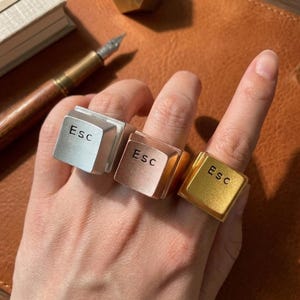 Puede incluir: Tres anillos en plata, oro rosa y oro, cada uno con la forma de una tecla "Esc" de un teclado. Los anillos se muestran en una mano, con el texto "Esc" visible en cada tecla. El fondo es una superficie similar al cuero.
