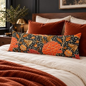 Pode incluir: Uma almofada lombar retangular longa com fundo preto e um padrão floral denso e intrincado em tons de laranja vibrante, vermelho profundo e dourado suave. A almofada está posicionada horizontalmente sobre uma cama com almofadas laranja sólidas.