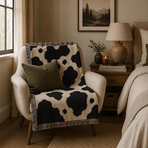 May include: A woven throw blanket featuring a black and cream cow print pattern with fringed edges, draped over a cream-colored upholstered armchair. A solid olive green square throw pillow rests on top of the blanket on the chair seat.