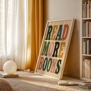 Puede incluir: Un cuadro rectangular enmarcado que presenta el texto READ MORE BOOKS en letras grandes, negritas y multicolores, incluyendo verde oscuro, naranja, amarillo y rosa, sobre un fondo crema claro, apoyado contra una pared.