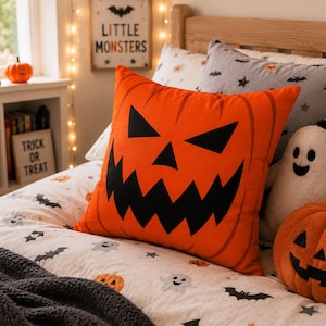 May include: A square orange throw pillow featuring a black jack-o'-lantern face with triangular eyes and a jagged mouth. The pillow is positioned on a bed with a white patterned duvet cover, a grey bat-print pillow, and a plush ghost toy.