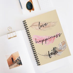 May include: A spiral-bound notebook with a beige cover featuring three illustrated feathers in shades of tan, pink, and brown. The words love, happiness, and faith are written in black cursive script across the feathers on the notebook cover.