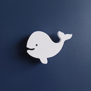 Può includere: Un pomello per cassetto a forma di balena bianca con una finitura opaca liscia e un piccolo dettaglio dell'occhio scuro. La balena è rivolta verso sinistra con una coda curva e una piccola pinna, montata su una superficie blu scuro.