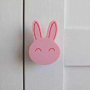 May include: A light pink cabinet knob shaped like a rabbit head with two long ears and two curved lines representing closed eyes. The knob is mounted on a white vertical panel with a visible seam running down the center of the surface.