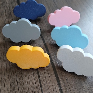 Op de afbeelding: Een verzameling van zes wolkvormige knoppen in diverse effen kleuren, waaronder donkerblauw, lichtroze, grijs, lichtblauw, geel en wit. Elke knop heeft een gladde, afgeronde silhouet met meerdere lobben voor bevestiging aan meubels.