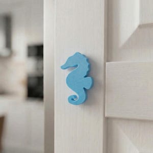 Könnte beinhalten: Ein hellblauer, seepferdchenförmiger Schrankknopf an einer weißen Kassettentür. Der Knopf hat eine matte Oberfläche und die ausgeprägte Silhouette eines Seepferdchens und dient als funktionaler Griff zum Öffnen und Schließen der Tür.