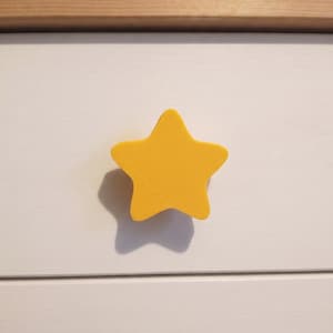 May include: A bright yellow, five-pointed star-shaped drawer knob with a smooth, matte finish. The star is mounted centrally on a flat, white, horizontal drawer front, casting a distinct shadow to its lower left side on the white surface.