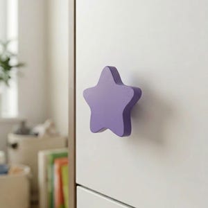 May include: A purple star-shaped cabinet knob with a smooth, matte finish attached to a plain white drawer front. The knob features five rounded points and a solid, uniform color, providing a simple handle for opening the furniture drawer.