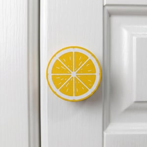 Op de afbeelding: Een ronde kastknop met een felgele en witte grafische print van een citroenschijfje. De knop is gemonteerd op een witte paneeldeur en toont een gedetailleerde dwarsdoorsnede van de citrusvrucht met segmenten en witte lijnen.
