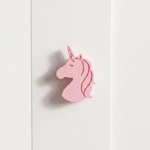 以下が含まれることがあります： 角と流れるたてがみを持つピンク色のユニコーンの頭のシルエット。ユニコーンは明るいピンク色で、白い背景に配置されています。画像はユニコーンの頭のクローズアップです。