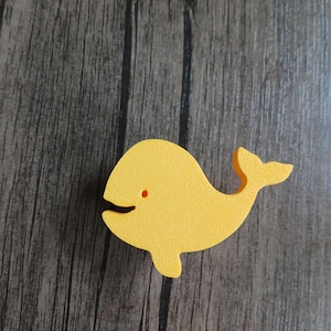 Puede incluir: Un pomo para mueble con forma de ballena de color amarillo brillante y superficie texturizada. La ballena presenta un corte sencillo para la boca y una pequeña hendidura circular para el ojo. Un tornillo metálico se sitúa aparte abajo.