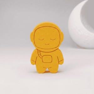 Può includere: Una statuetta di astronauta giallo brillante, solida e con superficie piatta, caratterizzata da una texture liscia e opaca. Il personaggio presenta un casco arrotondato con linee incise per gli occhi chiusi e un piccolo sorriso sul volto.