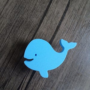 Pode incluir: Uma silhueta de baleia em espuma azul-clara com uma textura lisa e mate. A baleia apresenta uma forma corporal simples e arredondada, uma pequena barbatana caudal, uma barbatana peitoral e um pequeno recorte circular para o olho.