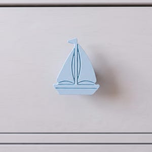 May include: A light blue drawer knob shaped like a sailboat with a single mast and two sails. The sailboat knob is mounted on a flat, light grey surface, featuring a smooth finish and a simple, clean silhouette for furniture hardware.