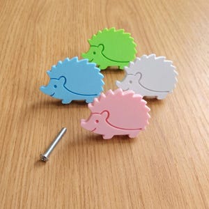 Puede incluir: Cuatro pomos para armario con forma de erizo en azul claro, verde lima, blanco y rosa suave. Cada pomo presenta una silueta sencilla y estilizada de un erizo con un acabado liso. Un tornillo de montaje de metal color plata se encuentra al lado.