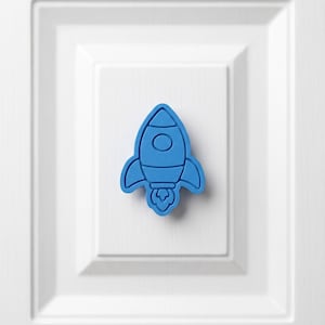 Peut inclure: Un emporte-pièce en forme de fusée bleu vif avec un contour défini, un détail de fenêtre circulaire au centre et des flammes stylisées émergeant de la base. L'objet présente un bord surélevé et une surface plane, montrant une forme moulée.