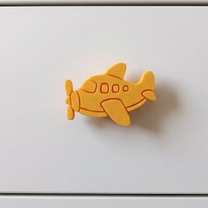 May include: A bright yellow airplane-shaped drawer knob with a matte finish. The design features a propeller at the front, a main body with three small rectangular windows, a wing, and a tail section. The knob is mounted on a flat surface.