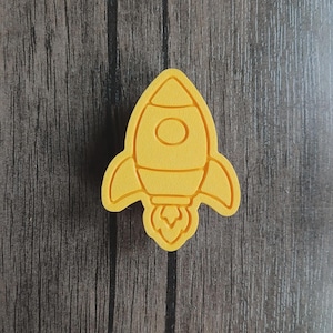 Puede incluir: Una forma de cohete de color amarillo brillante en tres dimensiones con una punta afilada, una ventana circular en el centro, dos aletas laterales y una llama estilizada en la base. El objeto tiene una superficie mate y texturizada.