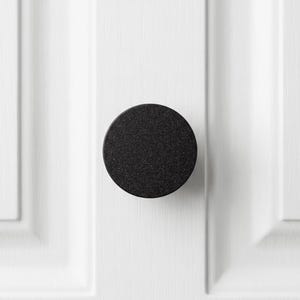 Puede incluir: Un pomo de armario circular con un acabado negro mate y texturizado. El pomo está montado sobre un panel de puerta de armario blanco que presenta detalles verticales empotrados, creando un contraste marcado entre el herraje y la superficie.