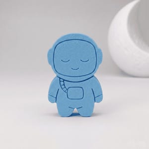 Può includere: Una figura di astronauta azzurro dalla forma piatta e dalla consistenza liscia e opaca. L'astronauta presenta un casco arrotondato, occhi chiusi, un piccolo sorriso e un semplice zainetto quadrato sul petto con dettaglio a tracolla.