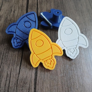 May include: Three rocket-shaped drawer knobs in blue, yellow, and white. Each knob features a raised rocket design with a circular window and exhaust flames. One blue knob is shown from the back, revealing a cylindrical base and a metal screw.