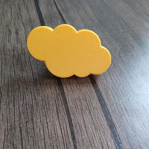 Puede incluir: Un objeto de color amarillo mostaza con forma de nube y acabado mate liso. El artículo presenta una silueta redondeada de varios lóbulos que imita la apariencia de una nube estilizada y esponjosa. La superficie es uniforme en color y textura.