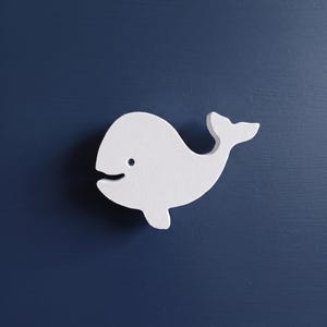 May include: A white whale-shaped drawer knob with a smooth matte finish and a small dark eye detail. The whale is depicted in a side profile view with a curved body, a small fin, and a tail fluke, mounted onto a solid dark navy blue surface.