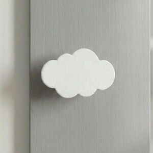 Puede incluir: Un pomo para mueble con forma de nube blanca y acabado mate. El pomo presenta un diseño de bordes redondeados y festoneados, montado sobre una superficie vertical de madera gris claro. El herraje es de color sólido y textura lisa.