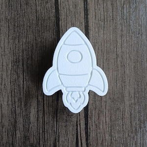 Può includere: Un oggetto bianco, piatto, a forma di razzo con una superficie testurizzata. Il design presenta un oblò circolare al centro, due pinne laterali e una fiamma stilizzata alla base, il tutto in un semplice stile a rilievo con contorni.