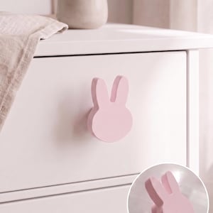 Puede incluir: Un tirador de mueble de color rosa claro con forma de cabeza de conejo y dos orejas erguidas. El tirador está montado en un frente de cajón blanco, y un recuadro circular muestra el mismo tirador con forma de conejo desde otro ángulo.