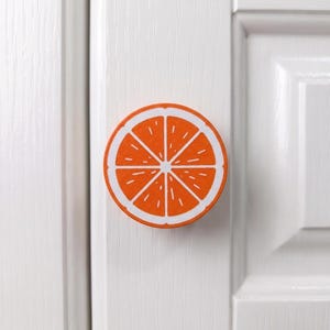 May include: A circular cabinet knob featuring a vibrant orange citrus slice design. The knob displays a bright orange centre with white segment lines and a white outer border, mounted on a white panelled door with a vertical wood grain texture.