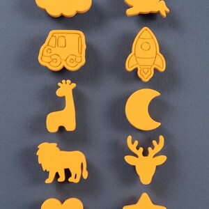 May include: A set of ten yellow, shaped objects arranged in two columns. The shapes include a cloud, a rabbit, a van, a rocket, a giraffe, a crescent moon, a lion, a deer head, a heart, and a star. Each piece has a smooth, matte finish.