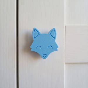 May include: A light blue cabinet knob shaped like a fox head with a smooth matte finish. The fox design features pointed ears, closed eyes, and a small triangular nose, all rendered in a simple, flat, and symmetrical graphic style.