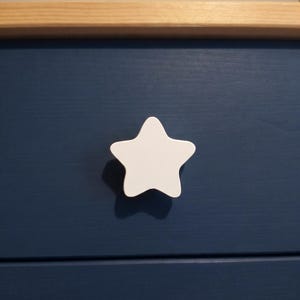 May include: A white star-shaped drawer knob mounted on a dark blue wooden drawer front. The drawer features a smooth, matte finish and is positioned directly below a horizontal light-colored natural wood trim piece at the top of the furniture.