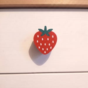 May include: A strawberry-shaped drawer knob featuring a bright red body with a subtle glitter finish, white teardrop-shaped seeds, and a dark green leafy top. The knob is mounted on a flat, white surface with a horizontal wooden trim at the top.