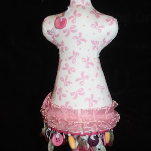 May include: A decorative dress form with a white fabric body adorned with pink bows. The form features a pink ruffled trim and is embellished with hanging buttons in various colors. The base is painted pink.