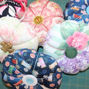 May include: A collection of handmade pincushions in various floral patterns and colors. The pincushions are shaped like pumpkins and feature decorative buttons and embellishments. The fabrics include pink, blue, and white floral prints, and a black and pink geometric pattern.
