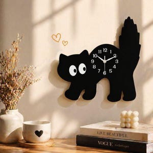 Puede incluir: Un reloj de pared negro con forma de gato con ojos blancos y una esfera blanca. El reloj tiene manecillas y números negros. El reloj está montado en una pared beige. Hay objetos decorativos en una superficie de madera.