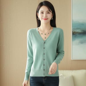 May include: A light sage green long-sleeve cardigan featuring a V-neckline and a vertical row of matching buttons down the center. The knit garment has a ribbed hem and ribbed cuffs, providing a clean, fitted silhouette that reaches the waistline.