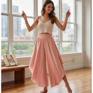 May include: A woman wearing a cream-colored knit crop top and flowing, wide-leg, blush-pink pants. The pants have a high waist and a flared design. The outfit is paired with delicate jewelry. The background includes a window and a wooden bench.