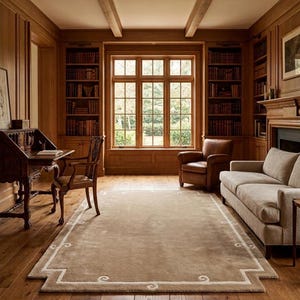 Op de afbeelding: Een gezellig bibliotheekinterieur met houten lambrisering, boekenkasten en een groot raam. Een beige tapijt met een decoratieve rand ligt op de houten vloer. De inrichting omvat een bureau, stoel, leren fauteuil en een bank.